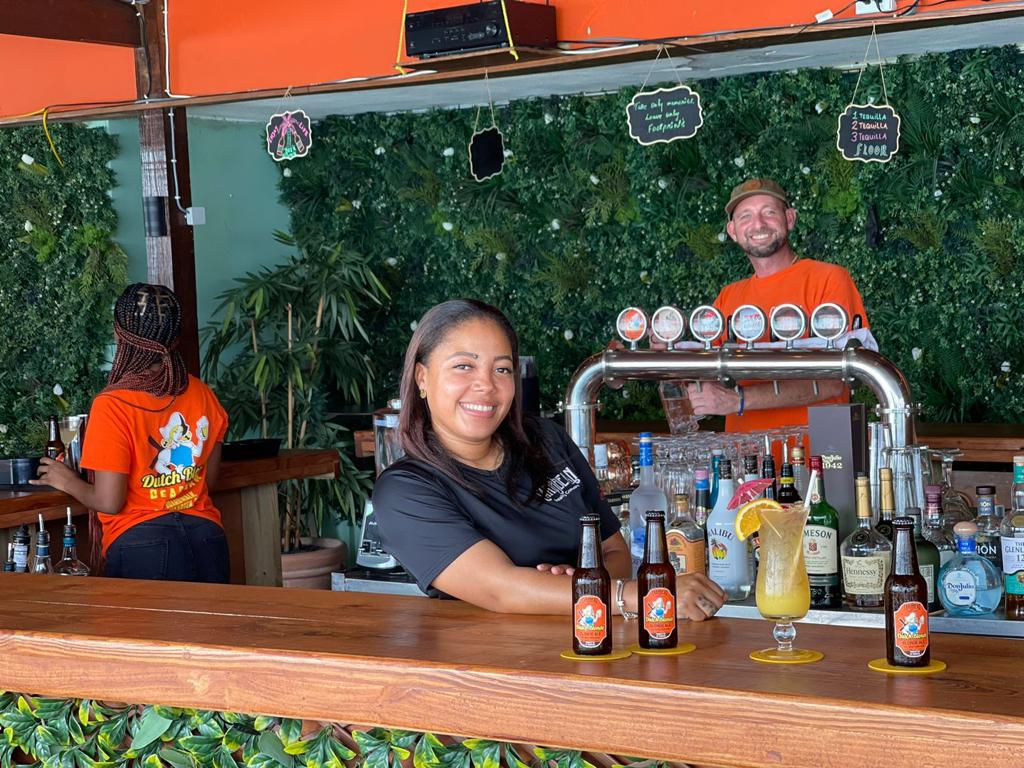 Woman smiling behind a bar in Dutch Blonde Beach Bar with three beers brewed on St Maarten / St Martin