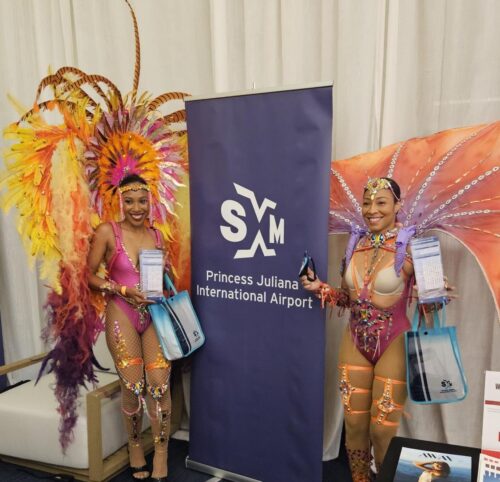 2 carnival girls next to a banner of SXM Airport at the SMART conference announcing the new airport schedule