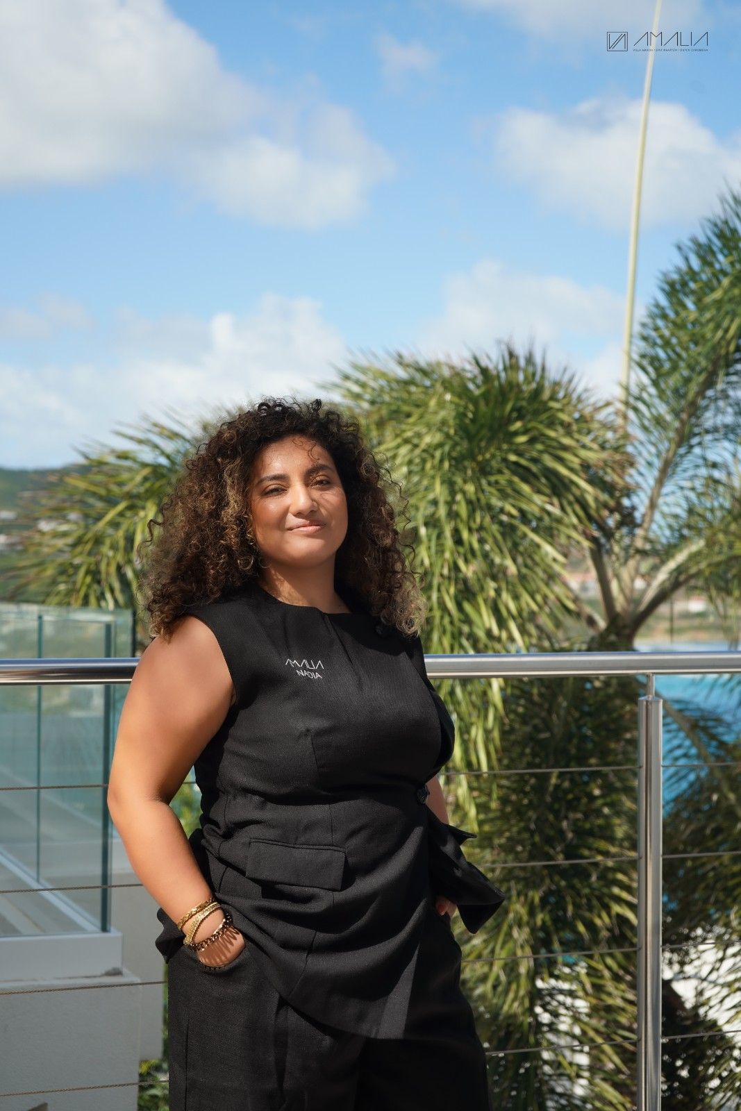 Nadia Ruhomutally posing on a balcony with a blue oacean and green trees behind here